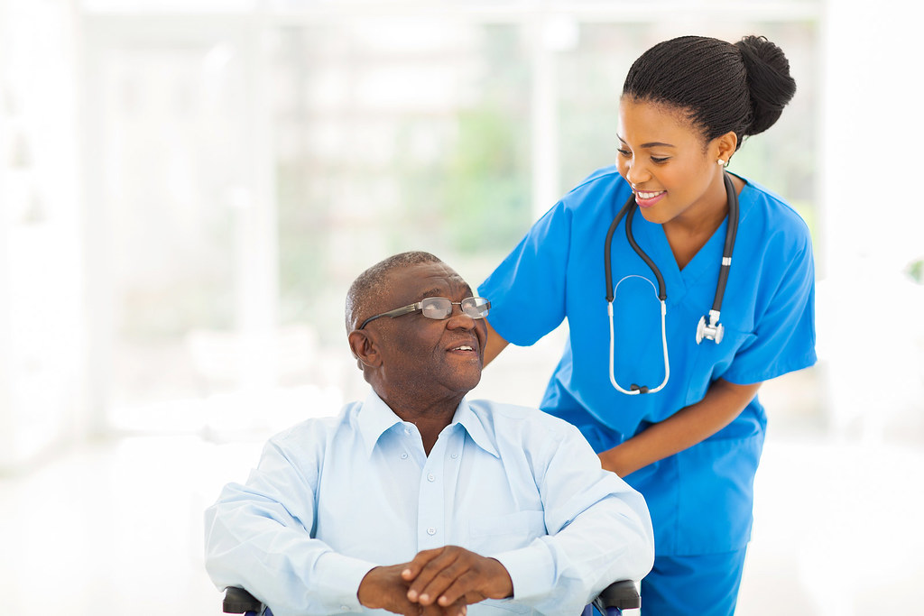A caregiver in blue scrubs assisting a smiling older veteran seated at home