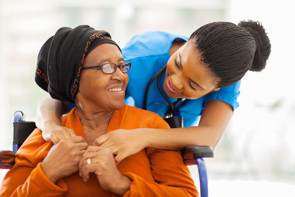 A caregiver leaning in to hold hands with a smiling senior woman she supports at home