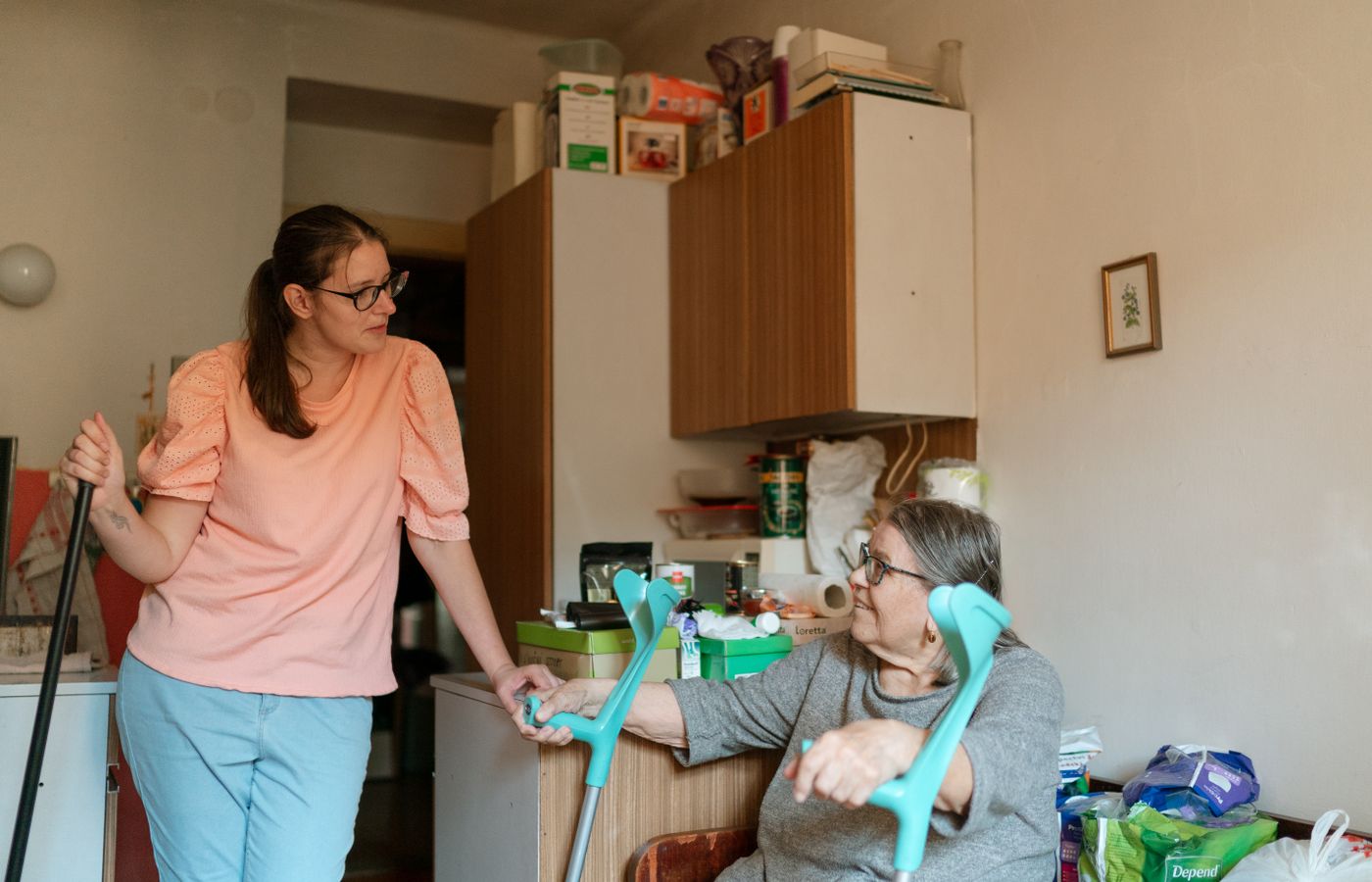 A caregiver tidying the kitchen while an older woman sits nearby